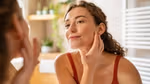 Woman in a bathroom, smiling at her reflection in the mirror. She has her hand to her face and is wearing a red top.
