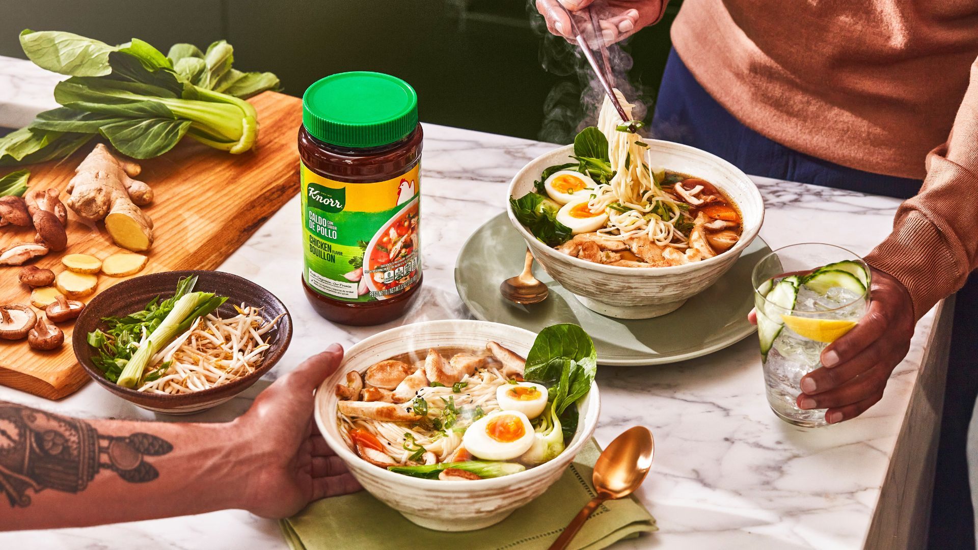 A table laid with two bowls of ramen noodles, a chopping board of ingredients and a jar of Knorr chicken bouillon.
