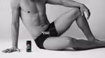 A bare-chested man in black pants sits on the floor in a relaxed pose, one knee raised with his wrist resting on it. A can of Lynx body spray is next to him. A “SCRATCH & SNIFF” label points to his shorts. The image is in black and white.