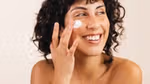 A woman with short dark curly hair smiles while applying a small dab of white face cream to their cheek with their fingers