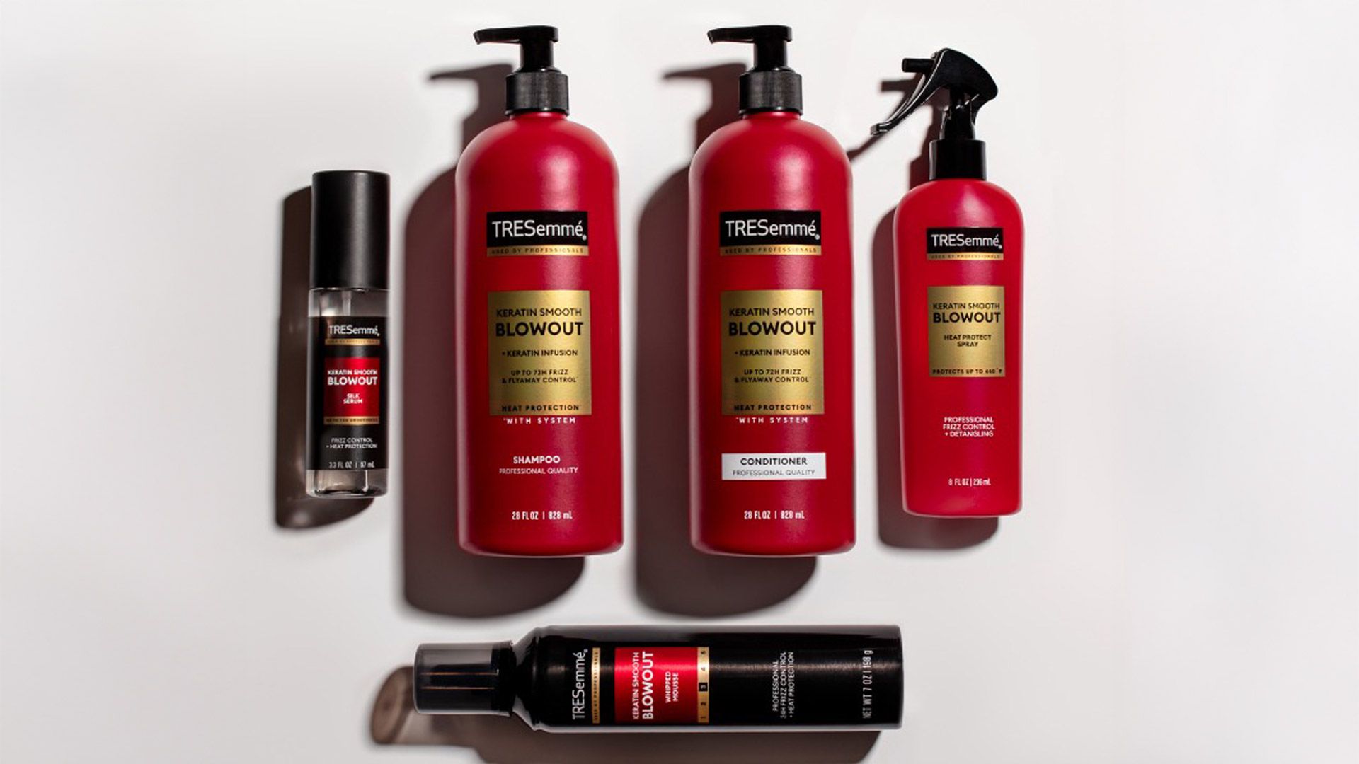 Five TRESemmé Keratin Smooth Blowout hair care products arranged on a white surface, including shampoo, conditioner, heat protection spray, styling mousse, and a small finishing serum. Packaging is a mix of red and black bottles with black caps and feature gold labels. The products are evenly spaced in a flat lay style with soft shadows.