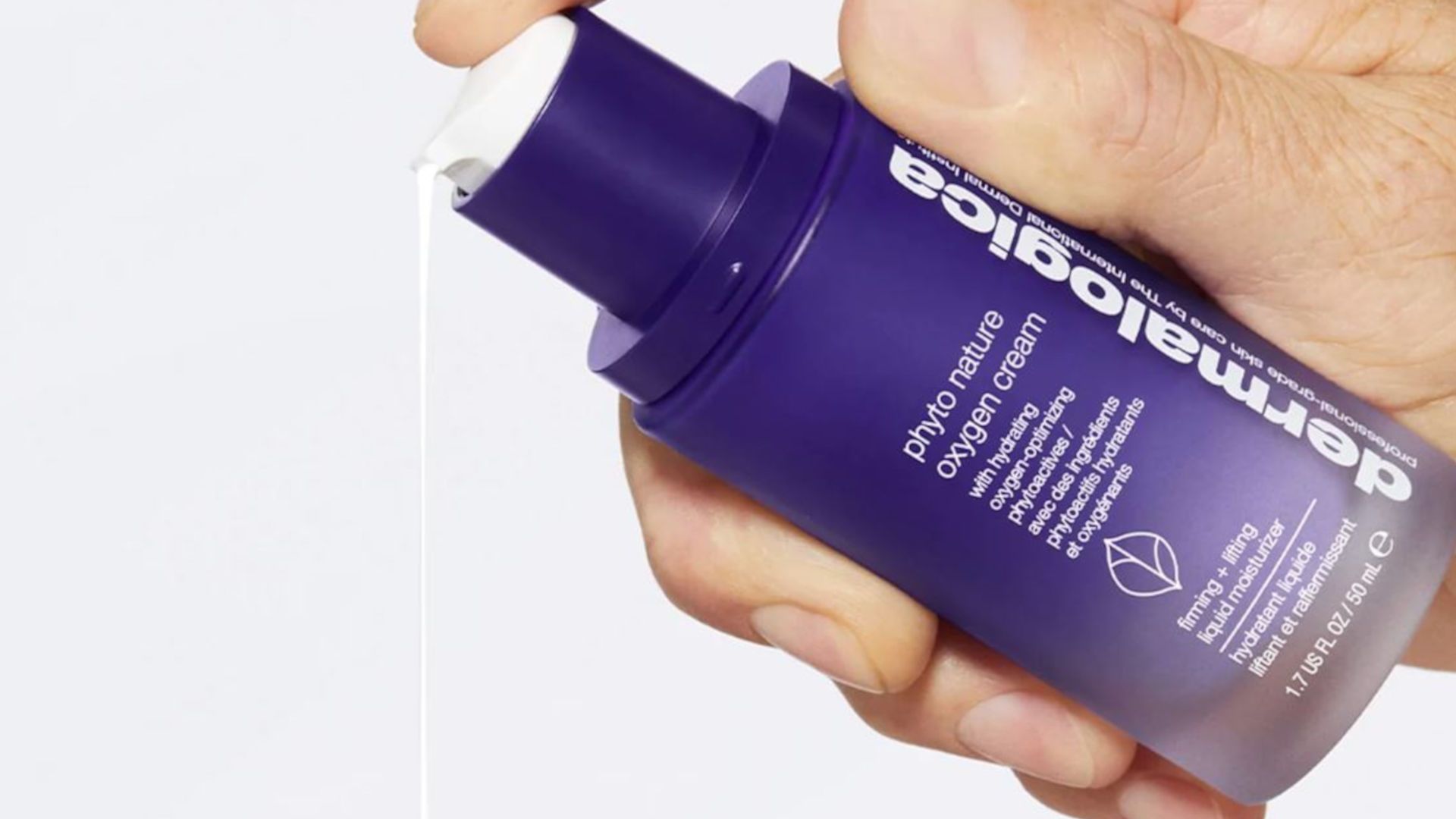 A close-up of a person’s hand holding a purple pump bottle of Dermalogica Phyto Nature Oxygen Cream. The hand is pressing down on the pump, releasing a thin stream of the white cream against a clean, light background. The bottle has a smooth, gradient finish transitioning from deep purple at the top to a lighter shade near the base, with white text detailing the product name and benefits. The overall scene is bright and minimal.