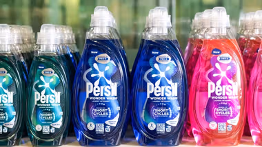 Persil Wonder Wash plastic laundry detergent bottles lined up in a row. There are three colours of detergent – green, blue and pink. The packaging labels explain that the product is designed for short cycles.